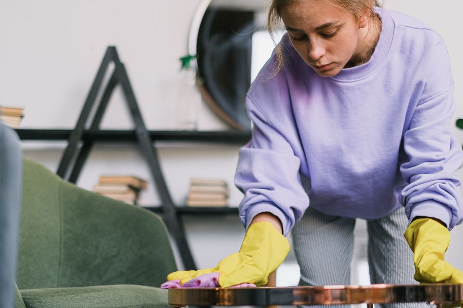 A young woman wearing a lavender sweatshirt and yellow cleaning gloves is disinfecting a small, round wooden table in a residential living room. The table appears polished and clean, with a reflective surface. In the background, there is a green upholstered armchair and a black metal shelving unit holding books and decorative items, indicating a well-maintained, modern interior. Natural light illuminates the scene, highlighting the tidy and hygienic environment. This image demonstrates surface cleaning in a domestic setting, emphasizing the importance of regular sanitisation for a clean and healthy living space, consistent with the services offered by Uxbridge Carpet Cleaning as part of home and domestic cleaning routines.