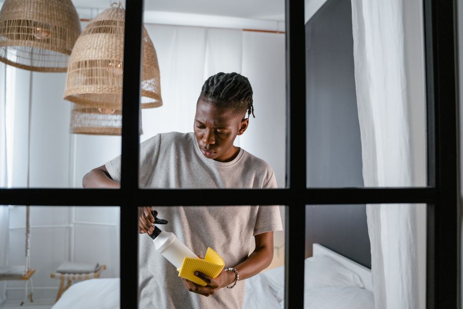A young woman with short braided hair, wearing a light-colored T-shirt, is performing surface cleaning in a modern bedroom. She is holding a spray bottle in one hand and a yellow cleaning cloth in the other, applying cleaning products to a surface. The room features a white bed with neatly arranged bedding, white tiled walls, and natural light entering through a window with white curtains. Hanging wicker pendant lamps provide ambient lighting, and the overall scene reflects a focus on domestic cleaning and maintaining hygiene. Uxbridge Carpet Cleaning, a professional cleaning company, specializes in deep cleaning and sanitisation services that help keep residential spaces like this bedroom clean and hygienic.