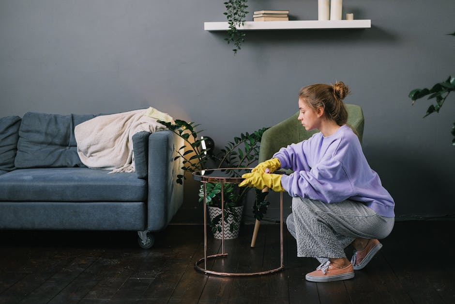 A young woman wearing a lavender sweatshirt and yellow cleaning gloves is disinfecting a small, round wooden table in a residential living room. The table appears polished and clean, with a reflective surface. In the background, there is a green upholstered armchair and a black metal shelving unit holding books and decorative items, indicating a well-maintained, modern interior. Natural light illuminates the scene, highlighting the tidy and hygienic environment. This image demonstrates surface cleaning in a domestic setting, emphasizing the importance of regular sanitisation for a clean and healthy living space, consistent with the services offered by Uxbridge Carpet Cleaning as part of home and domestic cleaning routines.