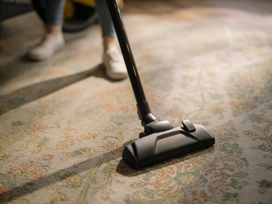 A close-up image of a person vacuuming a patterned, traditional-style area rug with a black and gray vacuum cleaner. The rug features intricate floral and geometric designs in muted shades of beige, green, and orange. The individual, wearing casual grey pants and white shoes, is standing behind the vacuum cleaner, which is positioned on the carpeted surface in a well-lit room. The scene illustrates surface cleaning aimed at maintaining hygiene and cleanliness, typical of services provided by Uxbridge Carpet Cleaning on Uxbridge High Street UB8.