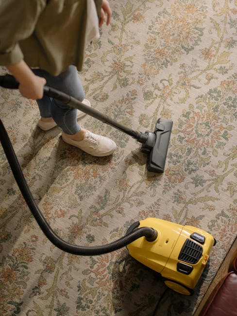 A person dressed in casual clothing and white sneakers is using a professional vacuum cleaner with a long hose attachment to deep clean a patterned area rug on a wooden floor in a home setting. The vacuum, branded Uxbridge Carpet Cleaning, is powered on, with the hose assembly positioned over the rug, which exhibits a floral design in muted tones. The room features soft lighting, and part of a leather armchair is visible in the corner, highlighting a tidy and well-maintained space. The image emphasizes domestic surface cleaning and the thorough removal of dust and dirt for hygiene and carpet maintenance, reflecting the professional cleaning services provided by Uxbridge Carpet Cleaning.