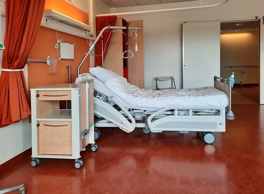 A hospital room featuring a white adjustable patient bed with side rails, positioned on a red vinyl floor. The bed has a white fitted sheet and a pillow, with medical equipment such as a blue suction container attached to the side rail. To the left, there is a mobile bedside table with a cream-colored drawer and a small cabinet on wheels, and an orange curtain partially drawn across a window with an air conditioning unit above. The background shows a white wall and an open doorway leading to a corridor. The room appears clean, with bright lighting reflecting off the surfaces, and Uxbridge Carpet Cleaning is focused on surface and deep cleaning for hygiene and maintenance, as promoted on the 'Hillingdon Hospital mattress deep cleaning services UB8, UXBRIDGE' page.
