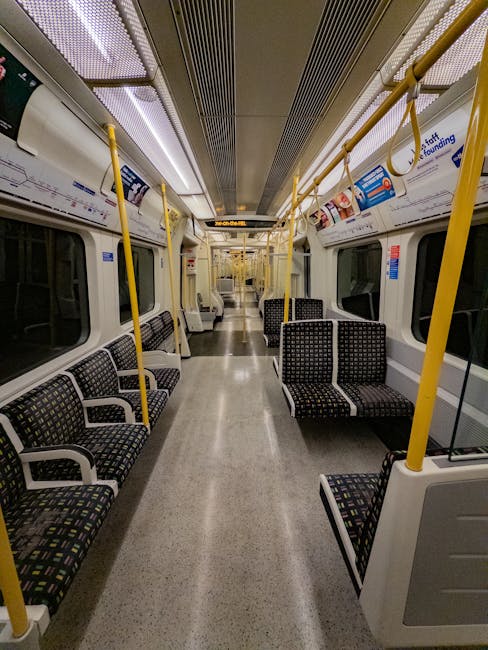 Interior of a Uxbridge Tube Station carriage showing an empty train with patterned fabric seats arranged in rows along both sides of the aisle. The carriage features large windows with closed blinds, yellow handrails hanging from the ceiling, and overhead advertising panels. The floor appears clean and polished, reflecting the overhead lighting, which illuminates a modern, well-maintained space. The ceiling has metallic vents and lighting strips running parallel, contributing to the overall tidy and hygienic appearance. Uxbridge Carpet Cleaning, a cleaning service provider, ensures the consistent hygiene and maintenance of such public transport surfaces through professional deep cleaning and sanitisation processes.