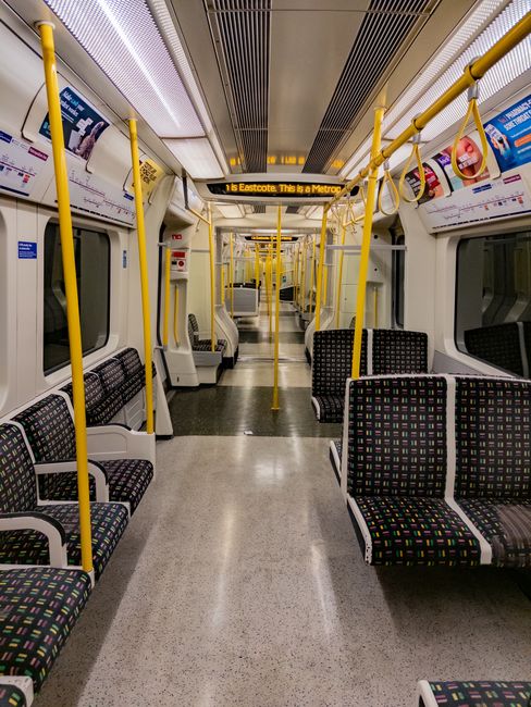 Interior view of a clean Uxbridge Tube train carriage, showing rows of patterned fabric-upholstered seats with black, red, green, and yellow geometric designs. The polished, light-grey flooring is free of dust or stains, and the white walls and ceiling reflect bright, even lighting from built-in fixtures. Yellow handrails run along the ceiling and vertically beside doors, which are closed and feature large glass windows. Overhead advertising panels display transportation information, and the carriage appears well-maintained with no visible litter or grime. This setting exemplifies surface cleaning and maintenance standards typical of professional commercial cleaning services, as provided by Uxbridge Carpet Cleaning for public transport environments, ensuring hygiene and safety for commuters.