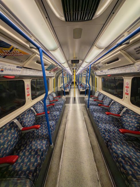 Interior of a Uxbridge Tube Station carriage showing an empty train with patterned fabric seats arranged in rows along both sides of the aisle. The carriage features large windows with closed blinds, yellow handrails hanging from the ceiling, and overhead advertising panels. The floor appears clean and polished, reflecting the overhead lighting, which illuminates a modern, well-maintained space. The ceiling has metallic vents and lighting strips running parallel, contributing to the overall tidy and hygienic appearance. Uxbridge Carpet Cleaning, a cleaning service provider, ensures the consistent hygiene and maintenance of such public transport surfaces through professional deep cleaning and sanitisation processes.
